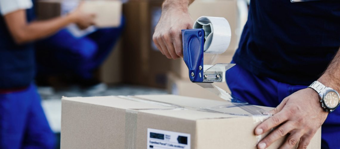 Close-up of delivery man closing carboard box with a tape while preparing packages for shipment.